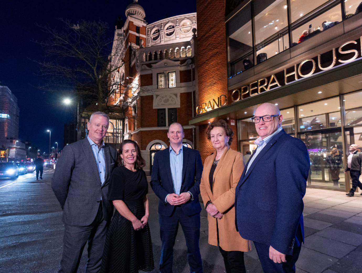 3 men and 2 women standing in a line in front of a building at night time. street lights and car lights are in the background.