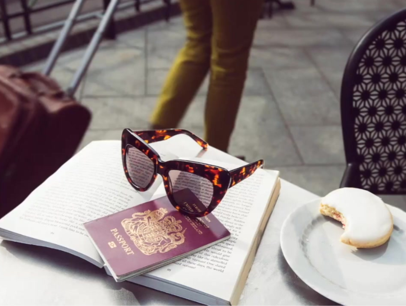 Photo of a pair of sunglasses, a passport, and a half eaten biscuit, on top of a book on a cafe table outdoors. In the background is someone walking pass with a suitcase.