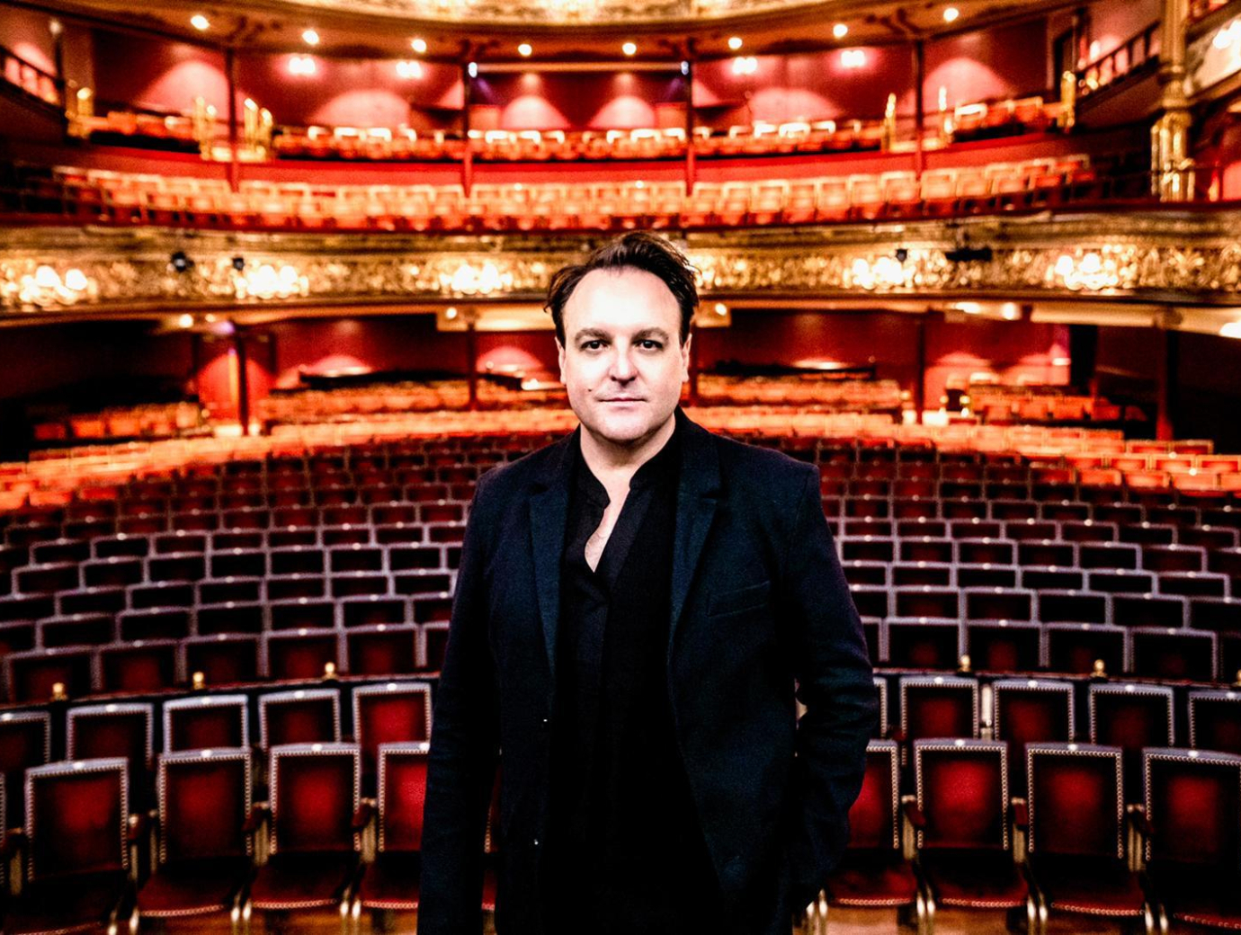 A man in a black shirt and suit on stage at the Grand Opera House, with lights on and theatre seating behind him.