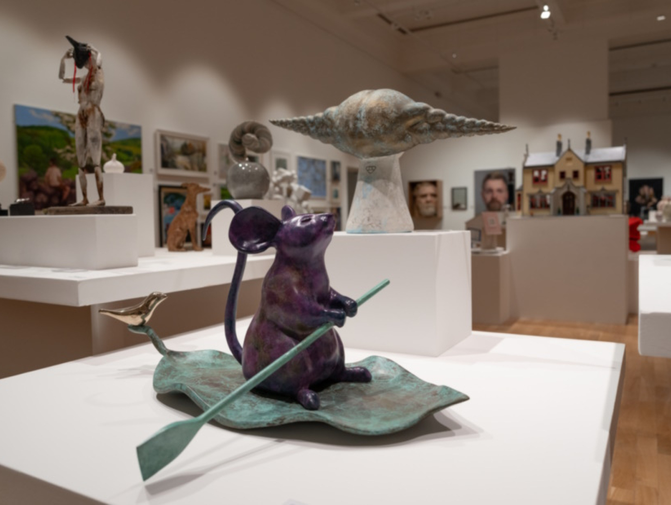 A bronze sculpture of a mouse on a leaf with a rowing stick sits on a plinth in the Ulster Museum gallery space.