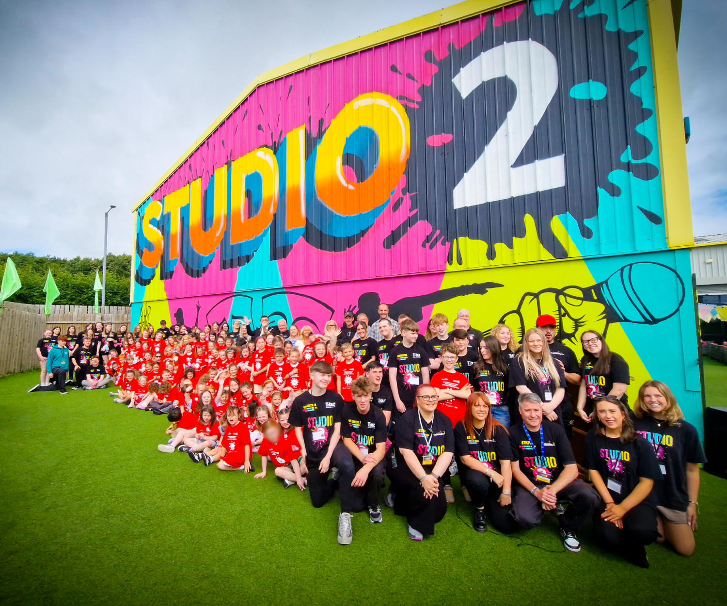 A large group of people pose in front of a building which has a colourful, vibrant mural with 'studio 2' painted boldly on it alongside a microphone, dancer and theatre masks.
