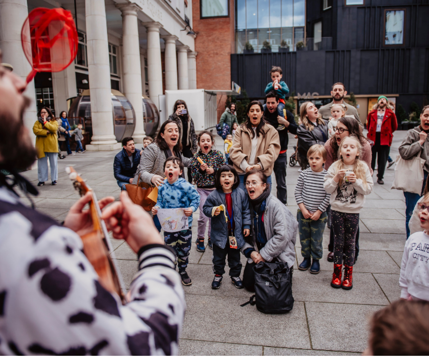 A group of young children outside with parents watching a street performer sing.