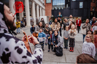 A group of young children outside with parents watching a street performer sing.