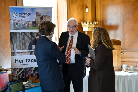 A man talking to two women with their backs to the camera, beside a Heritage Fund banner