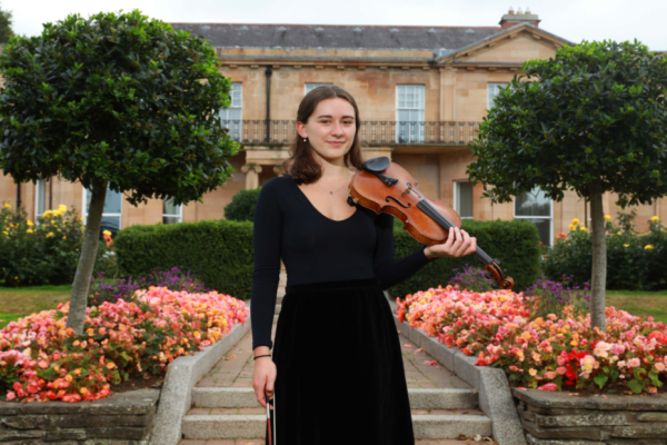Anna is standing outside with flower beds on either side of her.  She is wearing a long black dress and holding the violin.