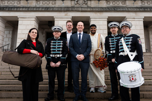 A woman and six men, some holding musical instruments, standing on the steps of Parliament Buildings at Stormont.