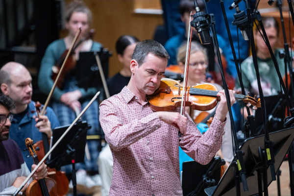 A man with short, dark hair, wearing a pink shirt, plays the violin on stage with an orchestra behind him.