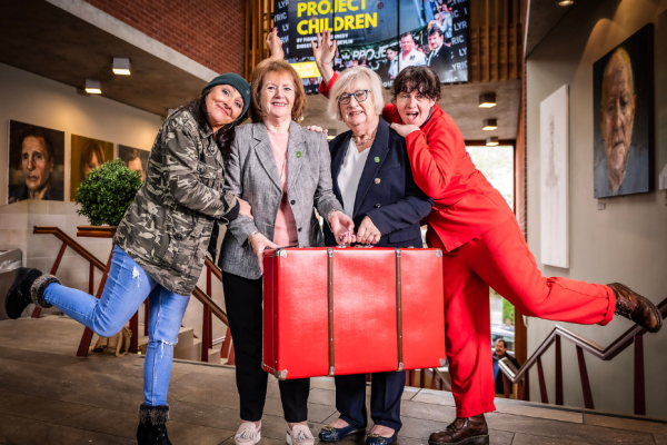 Four women standing in a theatre.  Two women in the middle are holding a large, red suitcase and the ones on outside left and right are each standing on one leg.