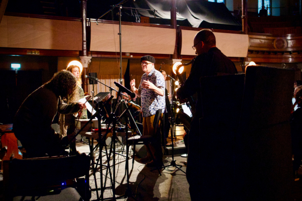 A man in a hat wearing glasses, conducting musicians in a studio setting with bright lights.