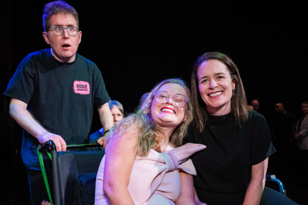Left to right - a man with short dark hair, wearing glasses and a t-shirt, standing behind a woman in a wheelchair with blonde hair and glasses, wearing a beautiful pink dress, sitting beside a woman with dark brown hair, wearing a black dress, smiling at the camera.