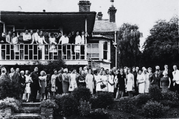A vintage photo in of the original Lyric Theatre building with a large group of people standing on the balcony and outside on ground level in the garden.