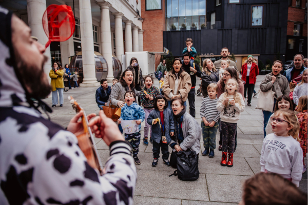 A group of children watching in excitement as a man dressed in a cow costume plays the Ukulele