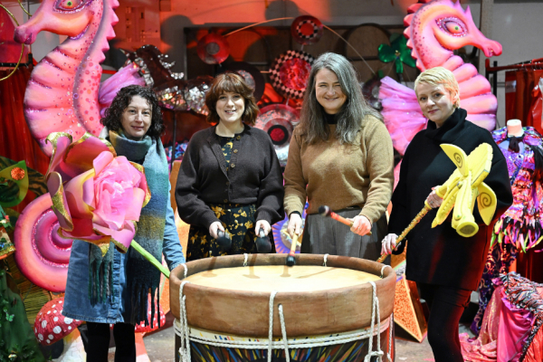 four women standing in front of a large drum holding arts props and smiling at the camera