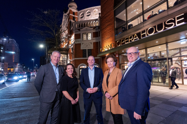 3 men and 2 women standing in a line in front of a building at night time. street lights and car lights are in the background.