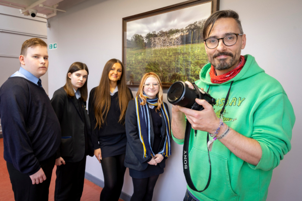 A group of sixth-form students standing with artist Robin Price beside his artwork which hangs on a wall inside the school.  The artist is holding a camera.