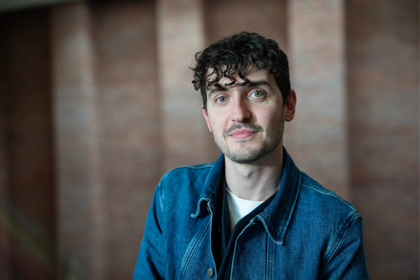 A portait of a man looking at the camera with dark brown hair wearing a denim shirt.