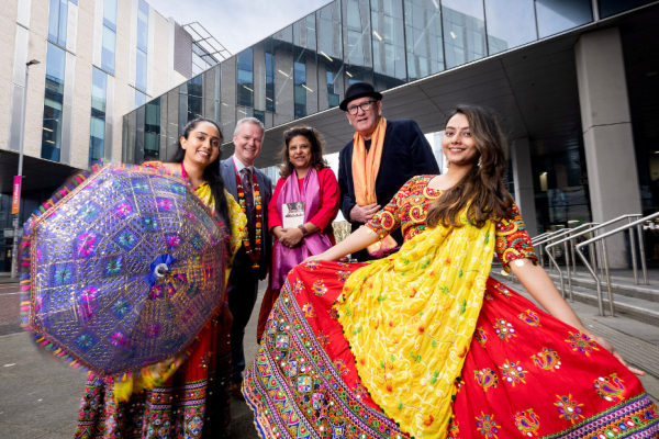 People standing outside Ulster University.  Indian dancers are wearing bright, colourful traditional costumes and holding a decorated colourful Indian umbrella.