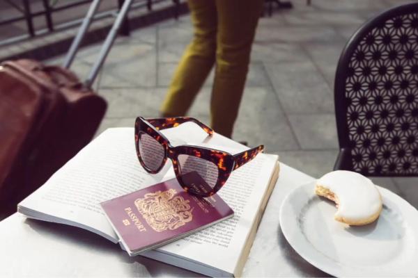Photo of a pair of sunglasses, a passport, and a half eaten biscuit, on top of a book on a cafe table outdoors. In the background is someone walking pass with a suitcase.