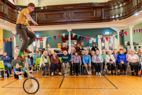 A man performing indoors on a unicycle in front of an audience of older people