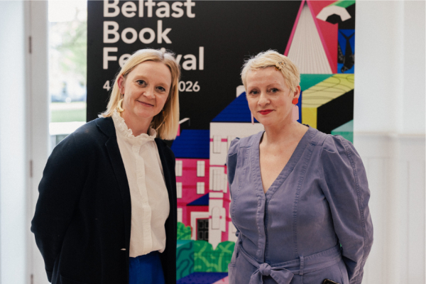 Two women standing in front of a banner and smiling at the camera. The banner reads 'Belfast Book Festival'.