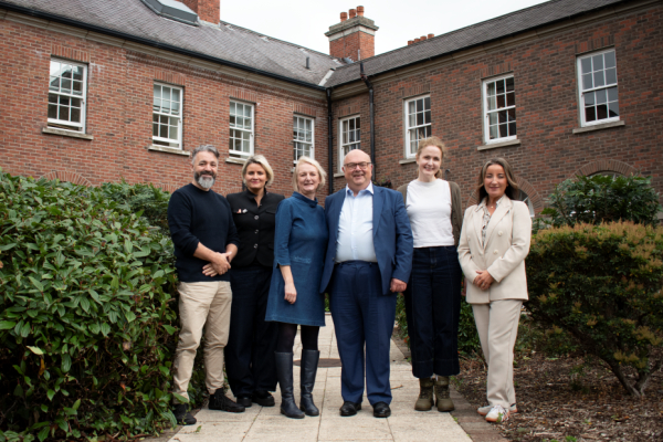 A group of six people standing outside in a line smiling at the camera with a red brick building in the background