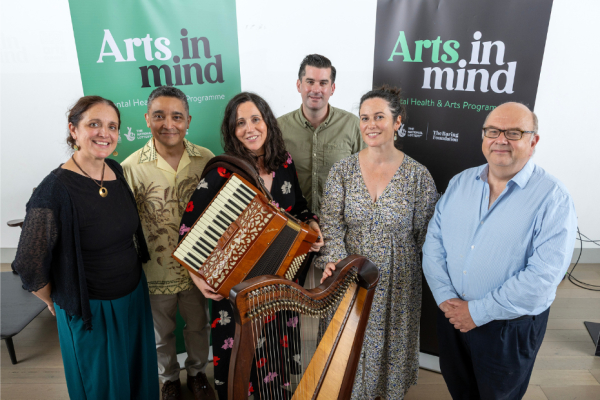 A group of three women and three men standing in front of two bannerstands which say Art in Mind. They are looking at the camera and smiling and the three women are holding instruments.