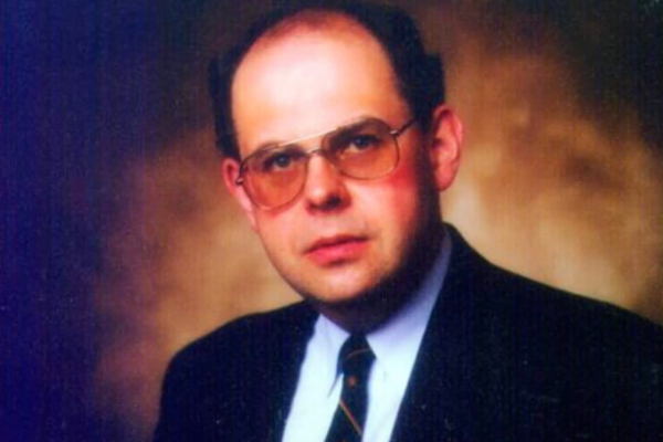 Male wearing glasses and wearing a dark suit with a white shirt and a dark coloured tie.