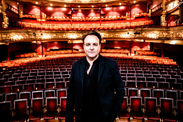 A man in a black shirt and suit on stage at the Grand Opera House, with lights on and theatre seating behind him.