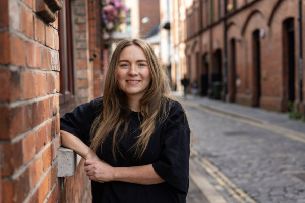 Claire Kieran standing outdoors against a red brick wall wearing a black t-shirt and smiling towards the camera.