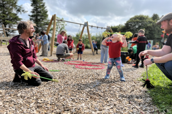 A young child throwing a stick in the air with a huge smile on their face.  The child is outside in a park area with swings in the background and people playing.