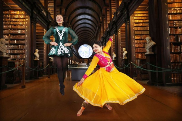 An Indian lady dancer in a bright yellow Indian dress, and an lady Irish dancer, with Trinity College Library in the background.
