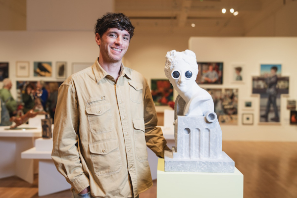 John is pictured in a gallery, artworks can be seen behind him. The white bust of a head has black goggles on its eyes
