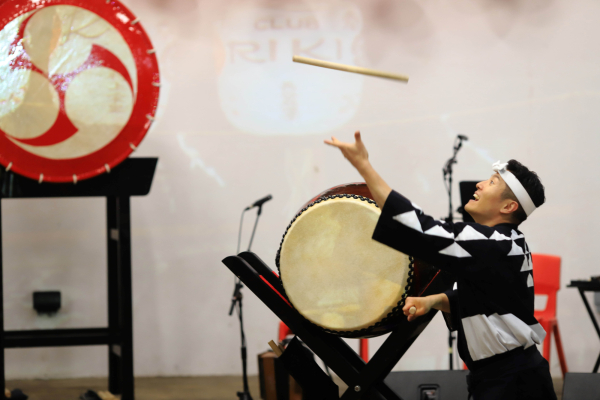 An image of a drummer, he throws a drum stick in the air, behind him are large Japanese drums