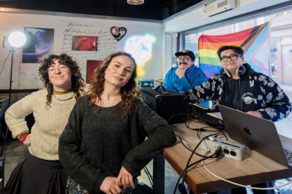 Four people standing beside musical equipment with an LGBTQI+ flag in the background.