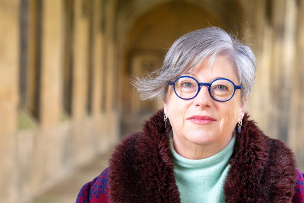 A woman with short grey hair, wearing glasses and a purple coat, facing the camera, with pillars of a university in the background.
