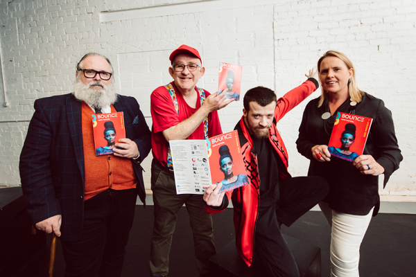 Three males and one female standing against a white stone background holding the printed programme for the 2022 Bounce! Festival.