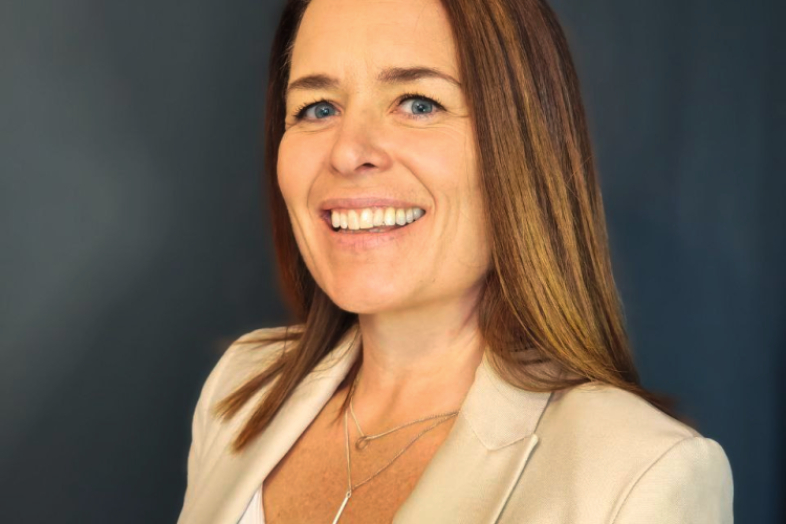 A woman with long brown hair wearing a white top and beige jacket.