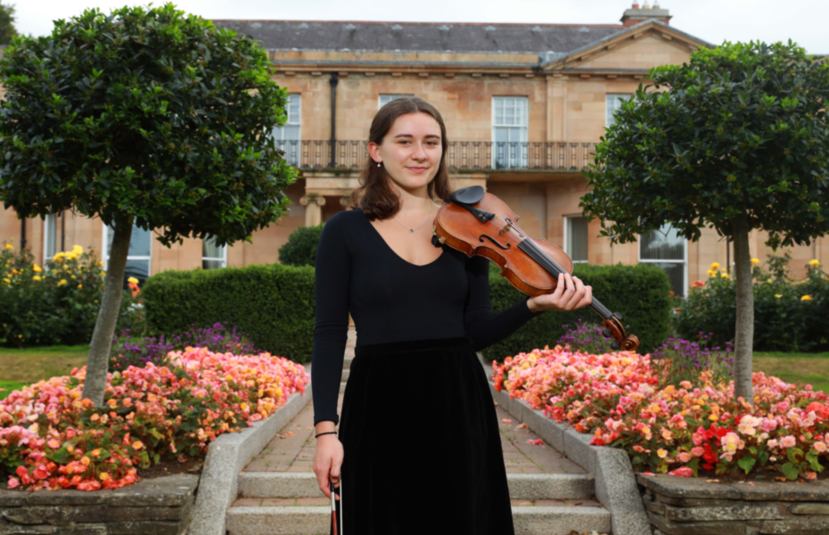 Anna is standing outside with flower beds on either side of her.  She is wearing a long black dress and holding the violin.