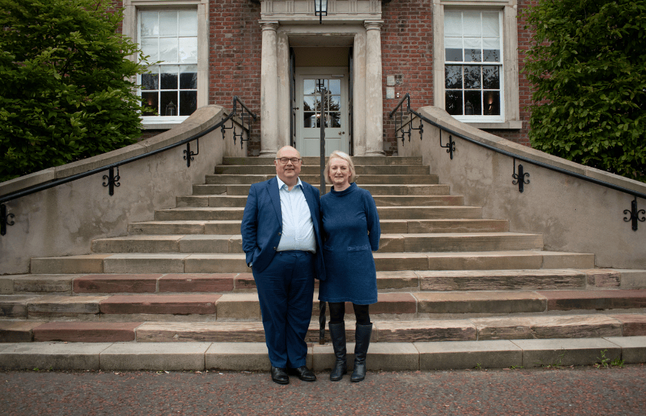 David and Lorraine stand side by side in front of steps outside. There are large windows and door in the background, alongside greenery on either side.