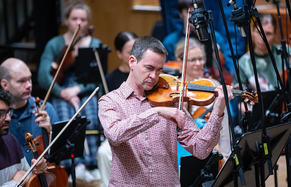 A man with short, dark hair, wearing a pink shirt, plays the violin on stage with an orchestra behind him.
