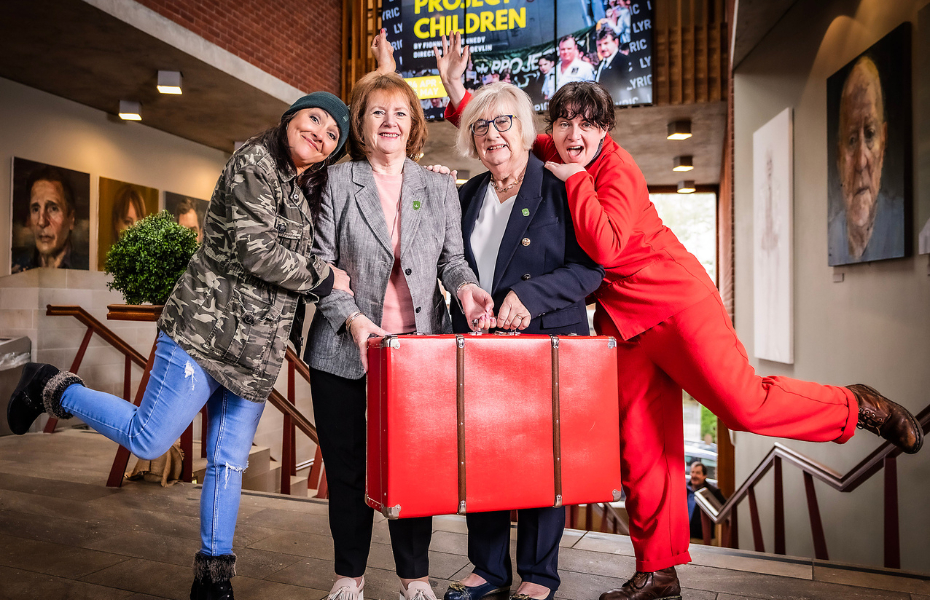 Four women standing in a theatre.  Two women in the middle are holding a large, red suitcase and the ones on outside left and right are each standing on one leg.