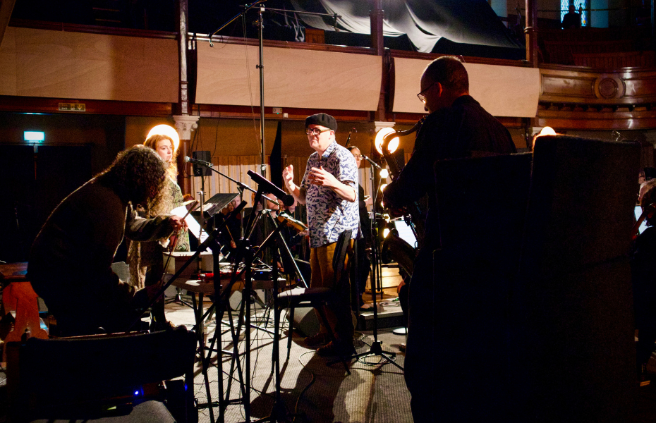 A man in a hat wearing glasses, conducting musicians in a studio setting with bright lights.