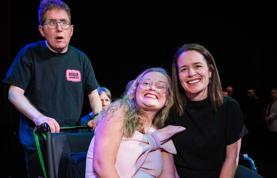 Left to right - a man with short dark hair, wearing glasses and a t-shirt, standing behind a woman in a wheelchair with blonde hair and glasses, wearing a beautiful pink dress, sitting beside a woman with dark brown hair, wearing a black dress, smiling at the camera.
