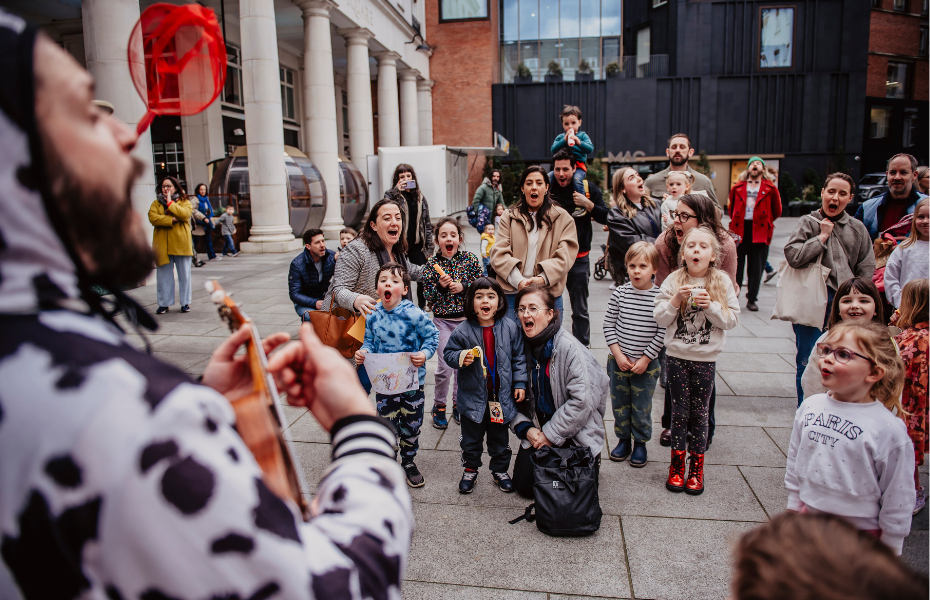 A group of children watching in excitement as a man dressed in a cow costume plays the Ukulele
