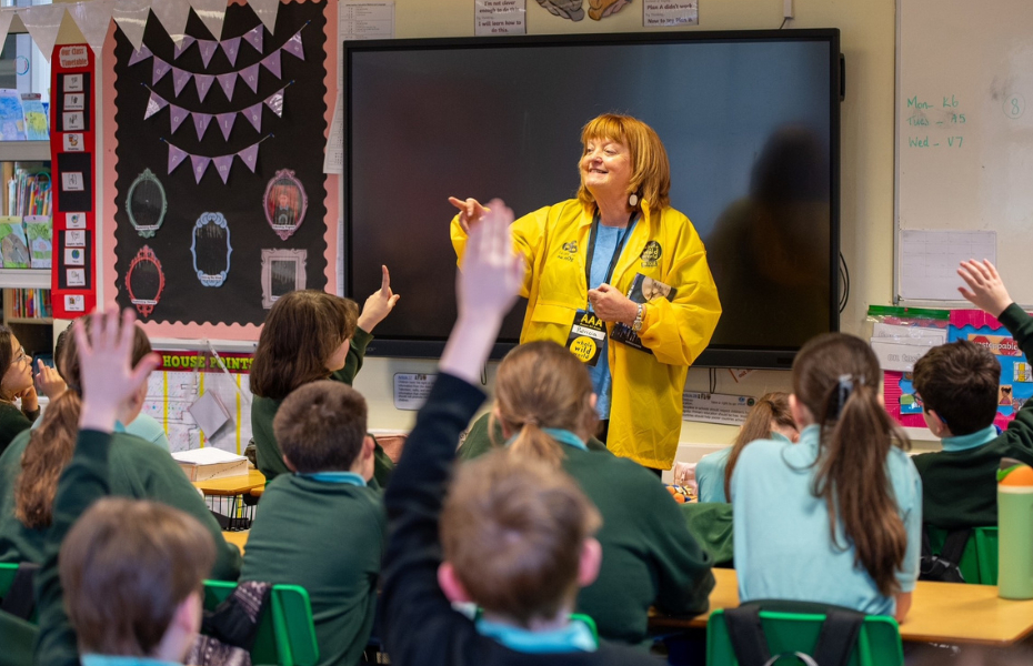 A woman standing in front of a classroom of pupils who are sitting with their backs to the camera and their hands up
