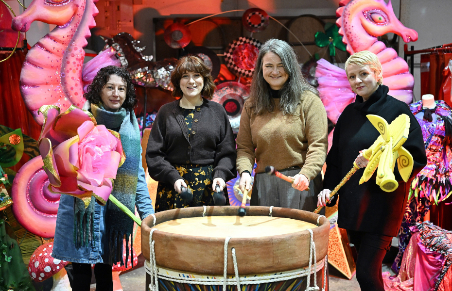 four women standing in front of a large drum holding arts props and smiling at the camera