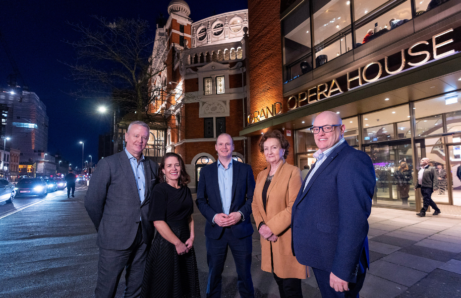 3 men and 2 women standing in a line in front of a building at night time. street lights and car lights are in the background.