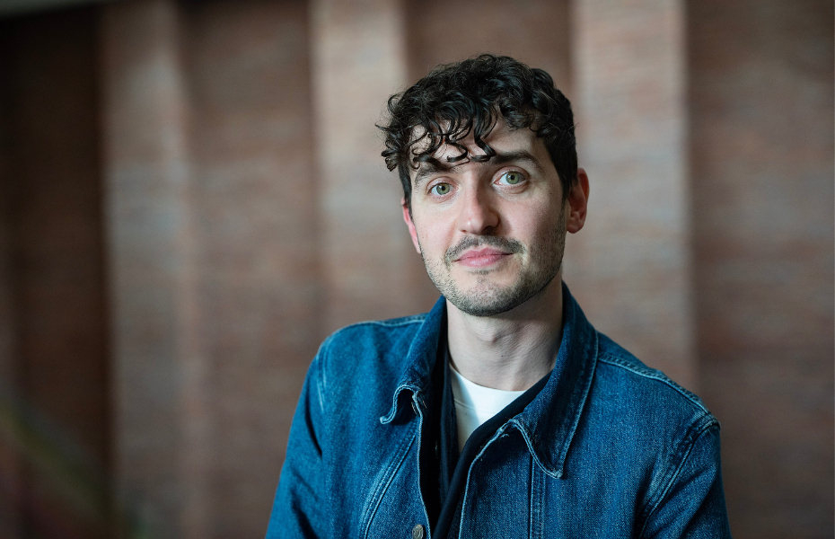 A portait of a man looking at the camera with dark brown hair wearing a denim shirt.