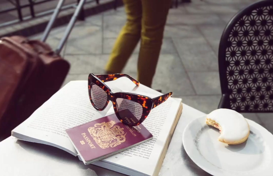 Photo of a pair of sunglasses, a passport, and a half eaten biscuit, on top of a book on a cafe table outdoors. In the background is someone walking pass with a suitcase.