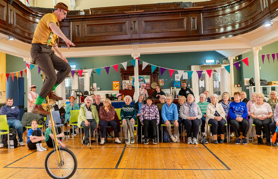 A man performing indoors on a unicycle in front of an audience of older people